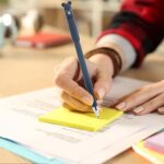 Person writing on a yellow sticky note with a pen featuring a bear design.