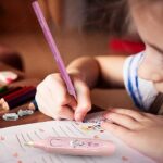 Child's hand holding a pink pencil over a piece of paper with a close-up of the pencil on a wooden surface.