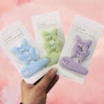 Three hair clips in blue, green, and purple packaging held by a hand against a pink background.