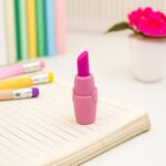 Pink lipstick-shaped eraser on a notebook with colorful pencils in the background