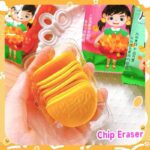 Yellow erasers in a clear plastic packaging held by a hand, with colorful packaging in the background.