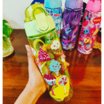 Colorful water bottle with cartoon designs held by a hand on a wooden surface.
