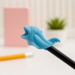 Blue dolphin-shaped pencil holder on a desk with a pink notebook and plant in the background.