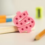 Pink textured object resembling a letter 'Q' on a desk with books and a pencil in the background