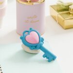 Blue key-shaped object with a pink heart on a light surface, with a pink container and gold gift box in the background.