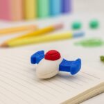 Small white eraser with blue handles on a notebook page with colorful pencils in the background