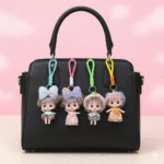 Cute Mini Doll Keychain for GirlsKeychainsCRAFTY CUDDLEBlack handbag with four colorful keychains shaped like cartoon characters on a pink backgroundPink Dress DollDollKeychain CuteAccessories GirlsAccessories BagCharm ReturnGiftIdeas KidsFashion KawaiiStyle PrincessTheme BirthdayReturnGift MiniDoll