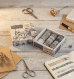 Decorative washi tape set in a matching box on a wooden surface with stationery items.