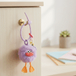 Cute Furry Luggage Tag KeychainKeychainsCRAFTY CUDDLEPurple plush toy with orange feet and a smiling face hanging on a wooden door handle.PurpleLuggageTag TravelAccessories CuteKeychain BagCharm TravelEssentials PomPomKeychain KawaiiAccessories SuitcaseTag GiftIdeas TrendingNow