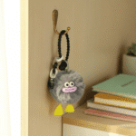 Cute Furry Luggage Tag KeychainKeychainsCRAFTY CUDDLEPlush keychain with a face attached to a hook on a wall next to books.GreyLuggageTag TravelAccessories CuteKeychain BagCharm TravelEssentials PomPomKeychain KawaiiAccessories SuitcaseTag GiftIdeas TrendingNow