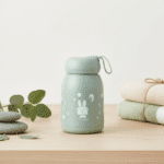 Leak Proof Rabbit Print Glass BottleCartoon Kids Water BottleCRAFTY CUDDLEGreen bottle with cartoon design on a wooden surface with folded clothes and leaves.Sage GreenKidsGlassBottle 350mlBottle GlassWaterBottle RabbitPrintBottle LeakProofBottle SiliconeCoverBottle ToddlerWaterBottle SchoolWaterBottle BPAFreeBottle ReusableBottle