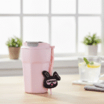 Cartoon Charm Insulated Coffee TumblerInsulated Coffee TumblerCRAFTY CUDDLEPink travel mug with a black and pink strap on a table with a glass of lemon water in the background.PinkCuteTumbler AestheticCups TrendyDrinkware PastelVibes KawaiiStyle ReusableCup HydrationGoals CuteAccessories GiftIdeas HomeEssentials