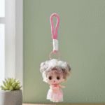 Plush keychain of a doll with a pink dress and white hat on a pink strap against a light green background.