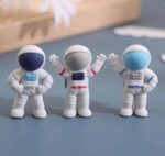 Three astronaut erasers with different colored helmets on a gray surface.