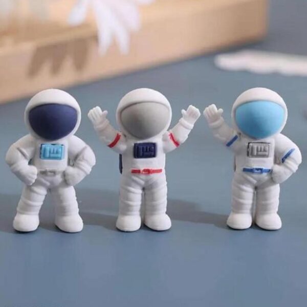 Three astronaut erasers with different colored helmets on a gray surface.