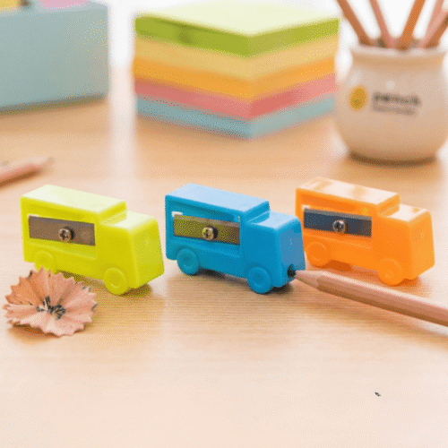 Three colorful pencil sharpeners shaped like cars on a wooden surface with pencils and paper in the background.
