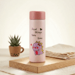 Kids Glass Water BottleCartoon Kids Water BottleCrafty CuddlePink thermos bottle with cartoon character and text on a wooden surface with a plant and folded fabric in the background.Pink