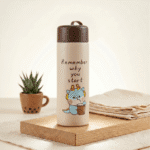 Kids Glass Water BottleCartoon Kids Water BottleCrafty CuddleBeige thermos with cartoon design and text on a wooden surface with a plant and folded towel.Brown