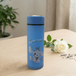Cartoon Print Glass Water BottleCartoon Kids Water BottleCrafty CuddleBlue thermos bottle with a cartoon dog design on a wooden surface with a white rose and green leaves.Sky BlueWaterBottle CuteBottle KidsBottle CartoonBottle GlassBottle SchoolEssentials BackToSchool KawaiiStyle HydrationGoals CuteAccessories