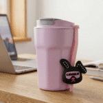 Pink travel mug with a cartoon dog tag on a desk next to a laptop