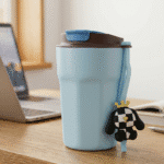 Light blue travel mug with a cartoon dog charm on a desk next to a laptop.